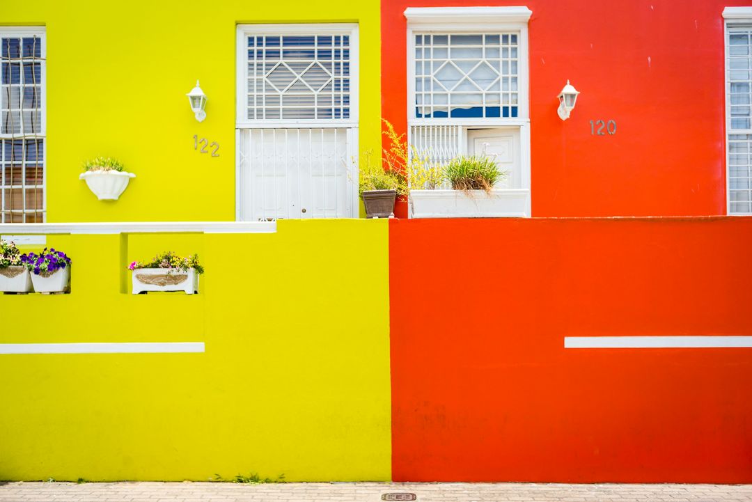 A yellow house and a red house side by side, white window frames and potted plants