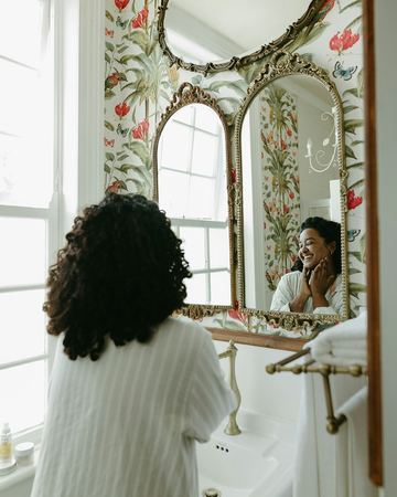 A guest in a white robe laughing into the gilded mirror of the floral bathroom