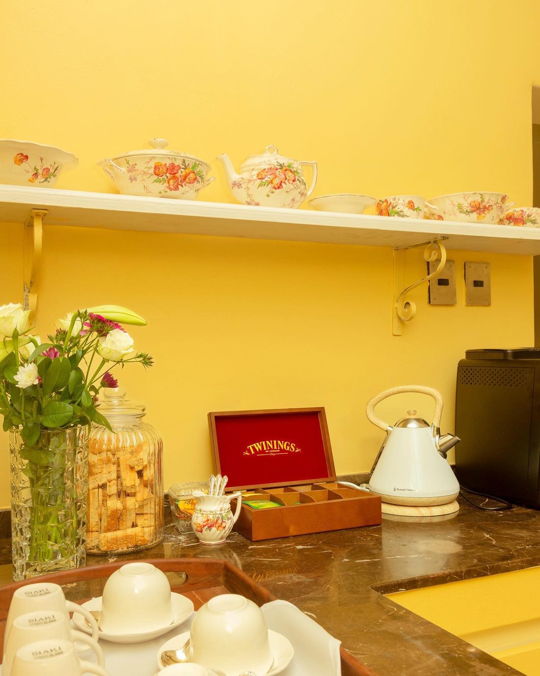 The tea station: fine china cups, a white kettle and a box of teas against a yellow wall